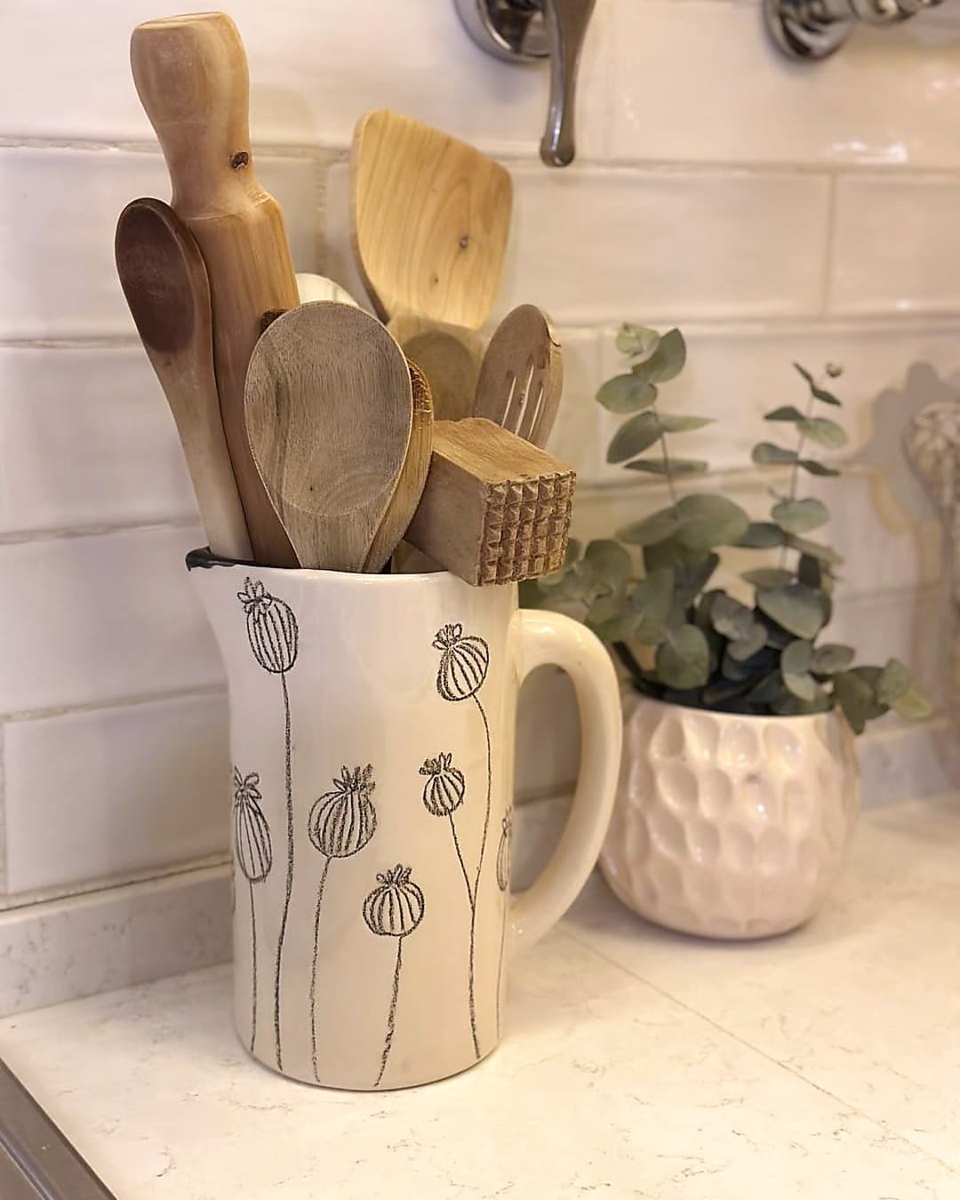 La Jarra Amapolas en la cocina de Cali, sobre la mesada con cucharas de madera adentro, junto a una planta de eucalipto