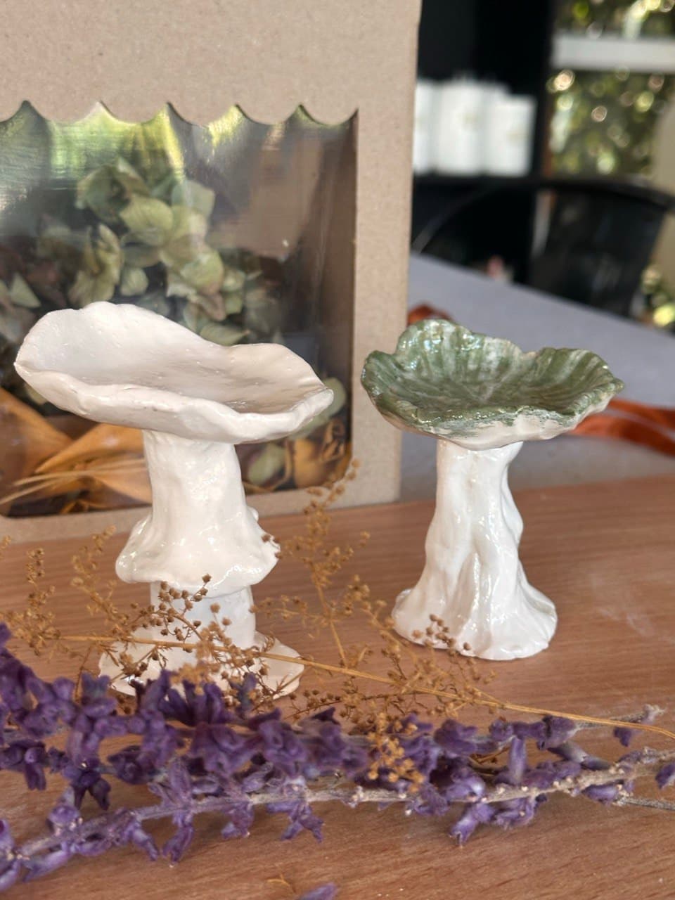 Dos hongos de cerámica modelados a mano sobre una mesa de madera, con ramitas de lavanda: uno todo blanco y otro con el sombrero verde y el tallo blanco