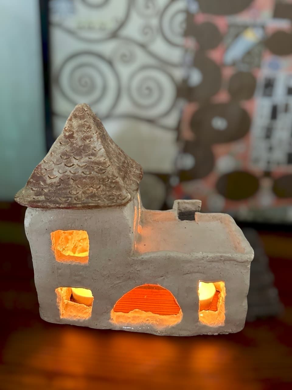 Casita de cerámica hecha a mano, con techo de tejas en relieve y una escalera al costado, iluminada por dentro con velas que brillan por las ventanas