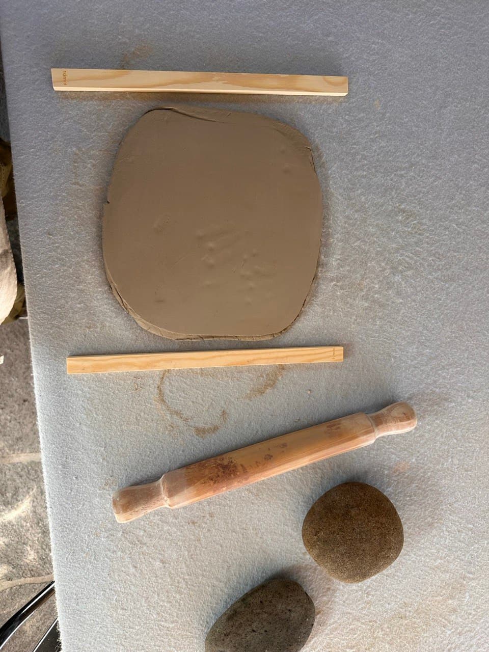 Una plancha de arcilla recién estirada entre dos listones de madera, con el palote y dos piedras al lado