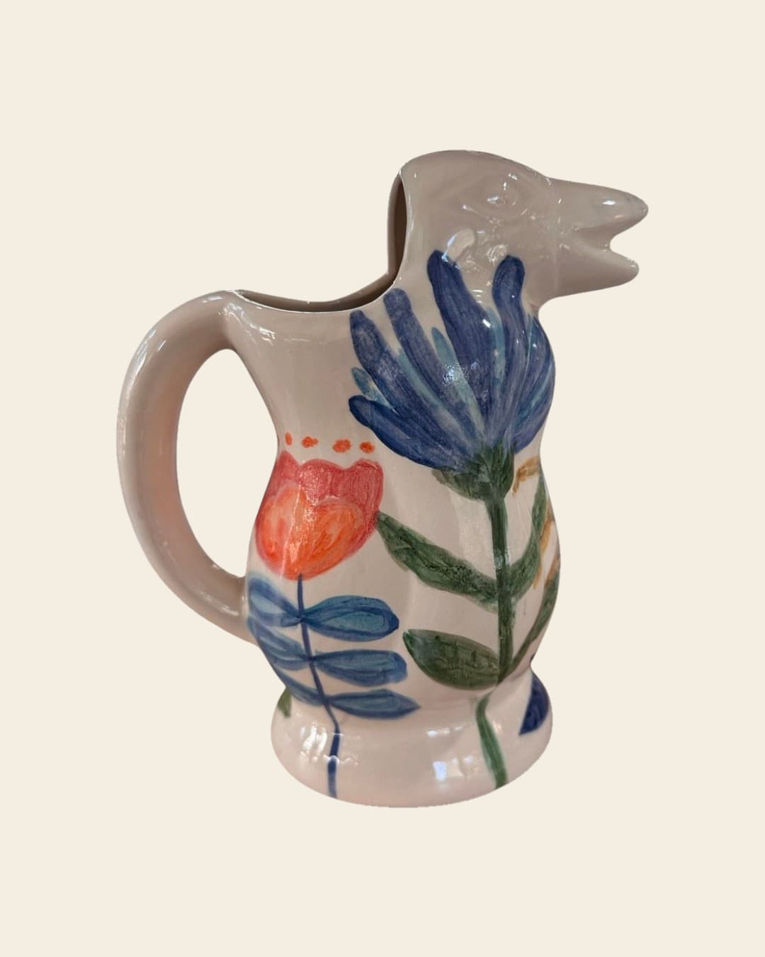 Jarra de cerámica color crema con el pico modelado como la cabeza de un pájaro, pintada a mano con una gran flor azul y un tulipán naranja de tallos verdes